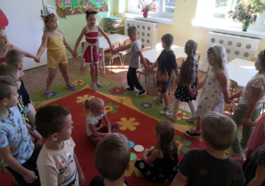 Przedszkolaki podaczas zabawy kołowej. W środku koła siedzące dwie dziewczynki z kolorowymi kropkami.