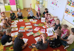 Grupa starszaków siedząca w kole. W środku koła leżą obrazki przedstawiąjące rożne rzeczy w kropki i prace plastyczne wykonane przez przedszkolaki.