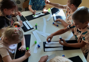 Dzieci przy stoliku wykonują pracę plastyczną - przejście dla pieszych. Naklejają białe pasy na czarny karton.
