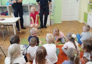 Spotkanie z policjantkami. dzieci siedzą na dywanie. Panie policjantki demonstrują jak należy prawidłowo siedzieć w foteliku samochodowym.