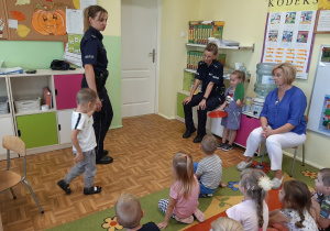 Panie policjantki w towarzystwie dzieci demonstrują zasady przechodzenia przez przejście dla pieszych z sygnalizatorem świetlnym.