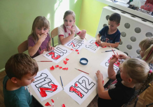 Dzieci siedząc przy stoliku wykonują pracę plastyczną - numer 112, techniką wydzieranki.