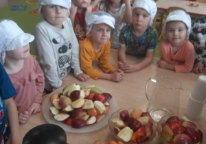 Przedszkolaki stoją w czapkach kucharskich przed obranymi warzywami i owocami