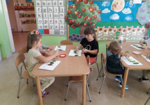 Przedszkolaki przy stolikach z zasuszonych liści tworzą jesienny obrazek.