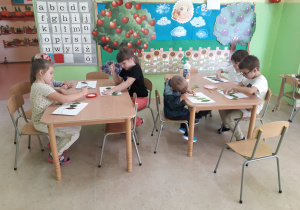 Przedszkolaki przy stolikach z zasuszonych liści tworzą jesienny obrazek.
