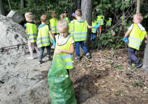 Przedszkolaki spacerują po lesie i zbierają śmieci.