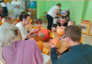 Dzieci wspólnie z rodzicami wycinają dyniowe minki.