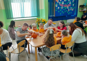 Dzieci wspólnie z rodzicami wycinają dyniowe minki.