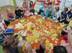 Grupa dzieci siedzi na dywanie w kole. na środku dywanu leżą jesienne liście.