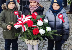 Dwóch chłopców i dziewczynka trzymająca kwiaty przed Urzędem Gminy