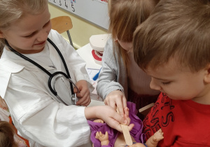 Scenka dramowa w grupie 5,6 latków. Dziewczynka - lekarz, bada lalkę którą trzyma dwójka innych dzieci.