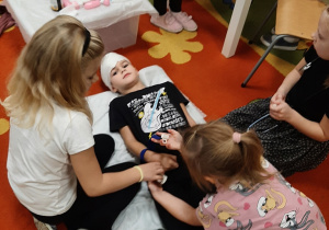 Scenka dramowa w grupie 5,6 latków dziewczynki za pomocą bandaża opatrują kolegę, który leży na leżance.