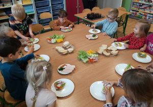 Dzieci z grupy 3,4,5 latków siedzą przy stole, na którym są zdrowe produkty. Przygotowują zdrowe i kolorowe kanapki, kolejne ujęcie.