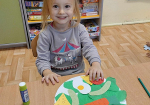 Kolejna dziewczynka z grupy 3,4,5 latków siedząc przy stoliku wykonuje pracę plastyczną pod tytułem "Zdrowy talerzyk".