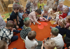 Dzieci siedzą w kole na dywanie, dziewczynka przebija pinezką papierowe serce.