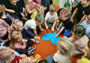 Dzieci siedzą w kole na dywanie, chłopiec przebija pinezką papierowe serce.