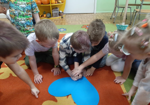 Kilku chłopców na dywanie, jeden z nich przebija papierowe serce pinezką.