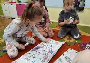 Kilka dziewczynek siedzi na dywanie dokleja kolorowe i błyszczące elementy ozdobne na list do Mikołaja - kolejne ujęcie.