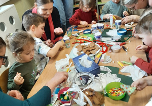 Dzieci i rodzice z grupy 3,4,5 latków przy stołach ozdabiają pierniki - następne ujęcie.