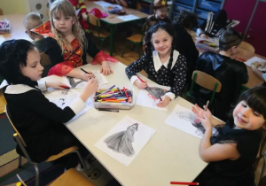 Dziewczynki przebrane za Wednesday siedzą przy stoliku i kolorują obrazki.