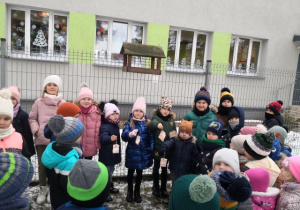Przedszkolaki stoją koło karmnika, w środku stoją dzieci i trzymają słoninę dla ptaszków.