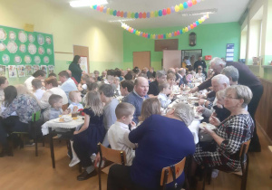 Babcie i dziadkowie siedzą przy stołach na holu przedszkolnym