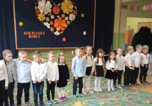 Dzieci stoją w rzędzie podczas występu. W tle dekoracja.