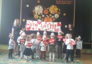 Dzieci młodsze stojąc w grupie śpiewają. W tle plakat z napisem "Sto-lat"