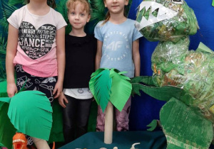 Dziewczynki pozują do zdjęcia w scenerii parku jurakskiego, obok stoi ogromny dinozaur wykonany z tektury i kolorowych gazet.