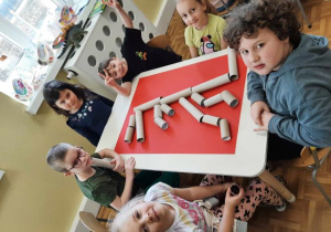Przedszkolaki siedzą przy stoliku i układają szkielet dinozaura z rolek.