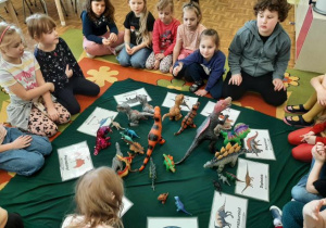 Przedszkolaki siedzą w kole, w środku leżą obrazki z dinozaurami oraz figurki, kolejne ujęcie.