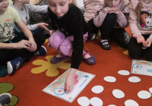 Dzieci siedzą w kole na dywanie, dziewczynka dopasowuje odpowiednie jajko do obrazka.
