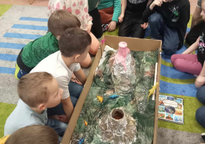 Dzieci siedzą na dywanie podczas eksperymentu. Na środku makieta z wulkanami i dinozaurami