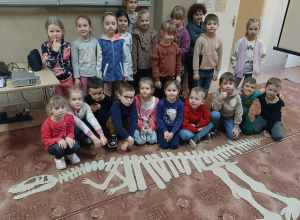 Dzieci w sali bibliotecznej pozują do zdjęcia. Na podłodze ułożony przez nie z tekturowych elementów szkielet dinozaura.