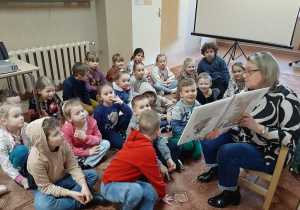 W bibliotece. Dzieci siedzą na dywanie, słuchają opowiadania czytanego przez Panią Bibliotekarkę.