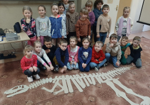 Dzieci w sali bibliotecznej pozują do zdjęcia. Na podłodze ułożony przez nie z tekturowych elementów szkielet dinozaura.