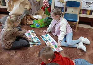 W bibliotece. Dzieci siedzą na dywanie i oglądają książki. W tle biblioteczny kącik dinozaurów.