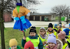 Dzieci ustawione w parach na placu przedszkolnym. Trzymają Marzannę.