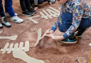 Dzieci składają na dywanie szkielet dinozaura dokładając poszczególne papierowe części