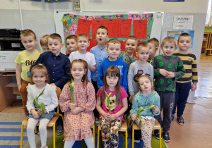 Dzieci z grupy 5-6 latków pozują do grupowego zdjęcia. Dziewczynki siedzą na krzesłach, a chłopcy stoją.