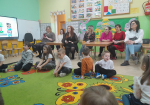 Dzieci na dywanie uczestniczą w zajęciu. Obok siedzą mamy i obserwują swoje dzieci.