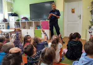 Dzieci siedą na dywanie, przed nimi stoi Pani policjantka. Prowadzi rozmowę na temat bezpieczeństwa.