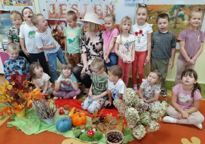 Dzieci pozują do fotografii z Panią Jesienią na tle gazetki tematycznej. Przed nimi jesienne dary: wrzos, kolorowe liście, dynia, kasztany.
