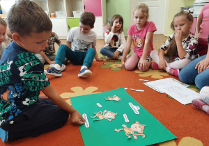 Dzieci siedzą na dywanie, chętne dziecko przykleja elementy na plakacie.