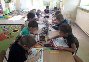 Dzieci przy stolikach kolorują plakat na Dzień Przedszkolaka.