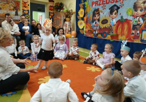 Przedszkolaki siedzą na dywanie i prezentują zabawę o wiewiórce. W środku koła podskakuje dziewczynka przebrana za wiewiórkę.