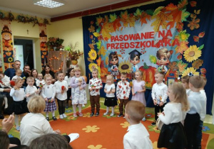 Grupa 3,4-latków stoi w półkolu na tle jesiennej dekoracji i grają na instrumentach.