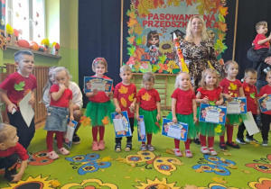 Dzieci w rzędzie trzymają dyplomy. Wychowawca grupy stoi za nimi.