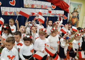 Dzieci stoją na tle dekoracji tematycznej o dniu niepodległości, trzymają w rękach flagi Polski.