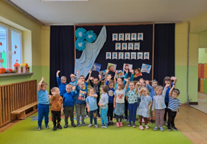 Dzieci na holu przedszkolnym pozują do zdjęcia grupowego z uniesionymi rączkami.